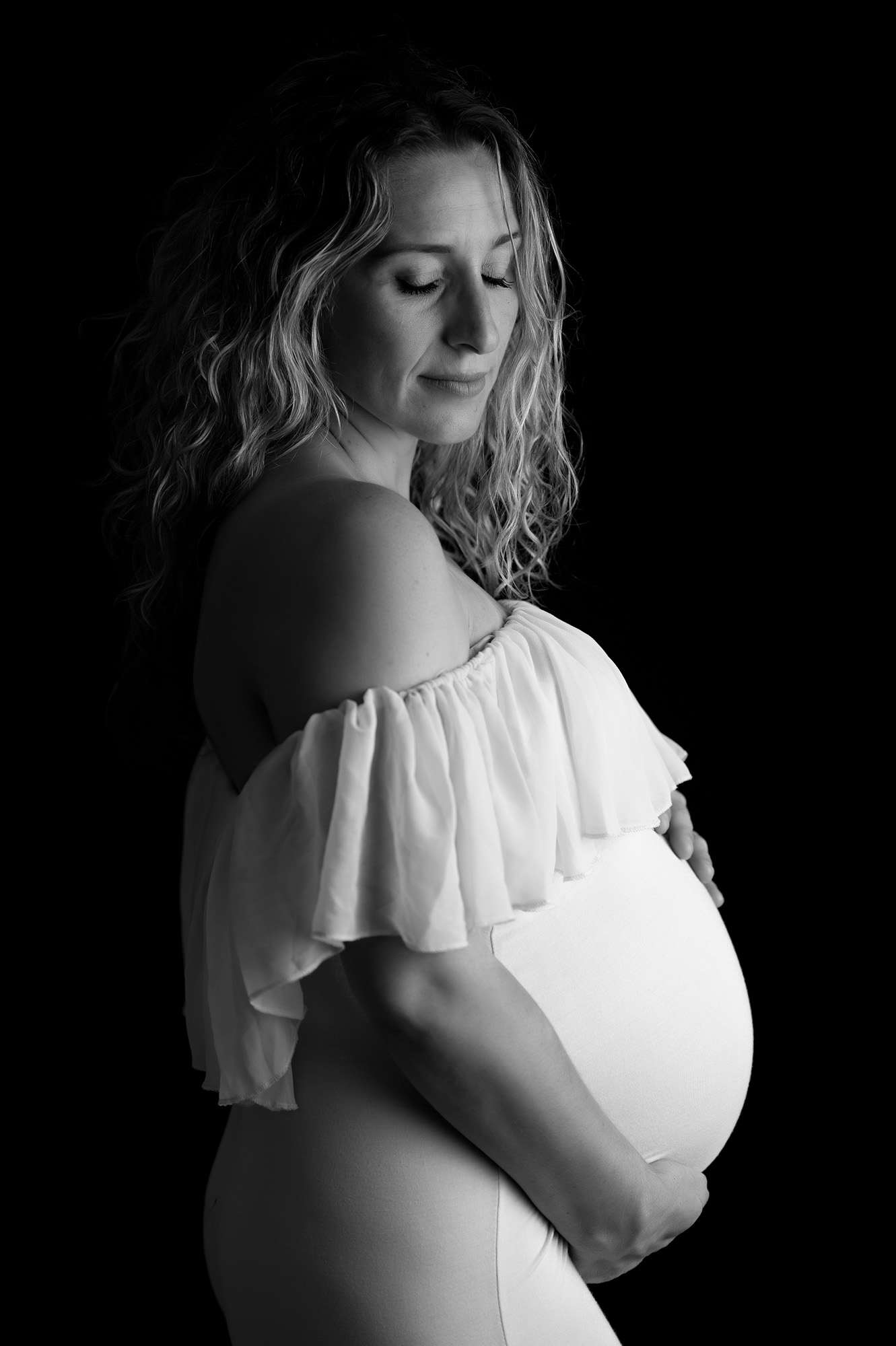 photo noir et blanc d'une femme enceinte habillée d'une robe avec un volant au niveau de la poitrine. Son regard est incliné vers le bas et elle a les yeux fermés.