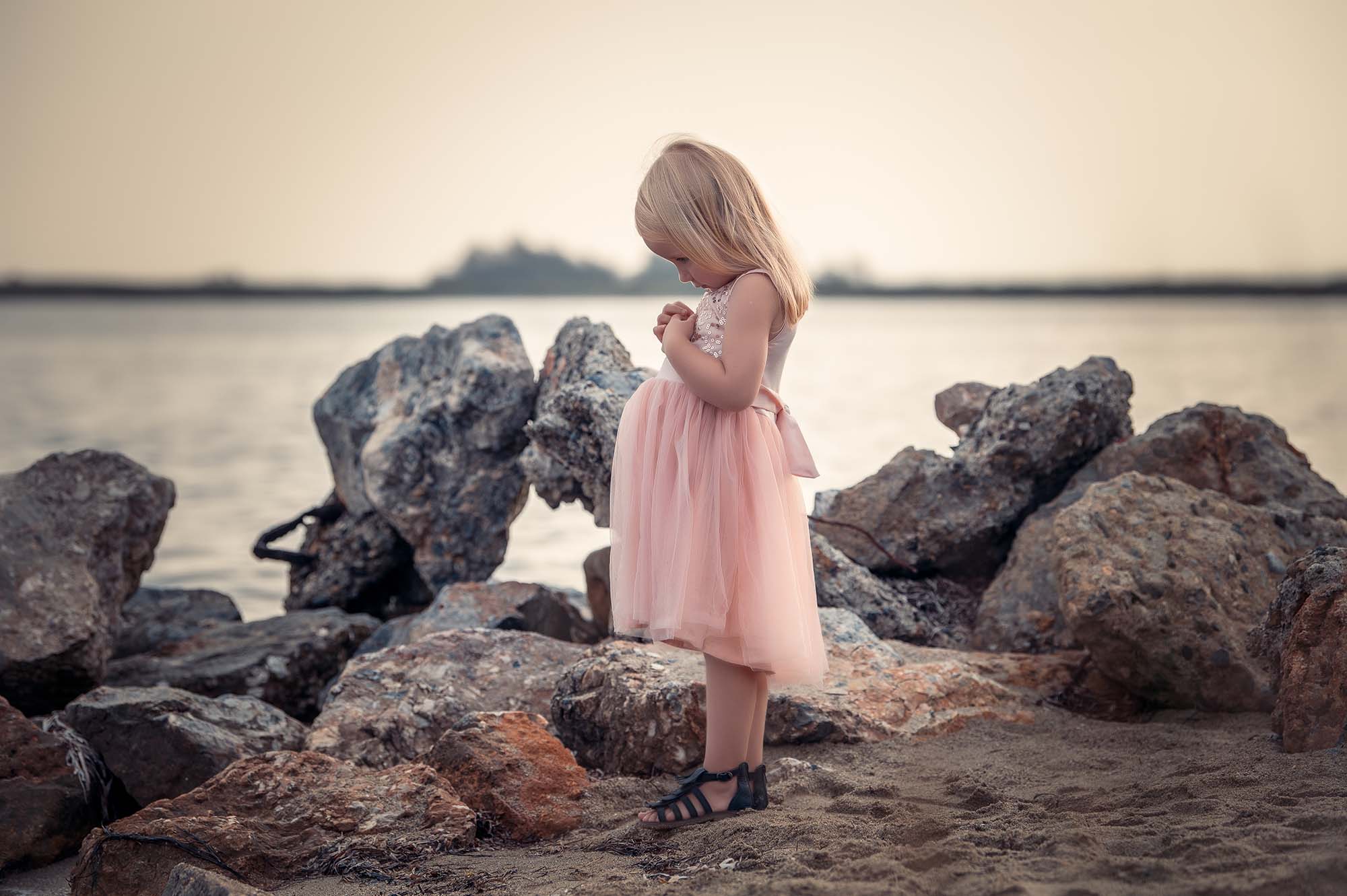 petite fille de profil qui fait la tête en se tenant les mains, elle est habillée en robe rose , derrière elle il y a des rochers et de l'eau