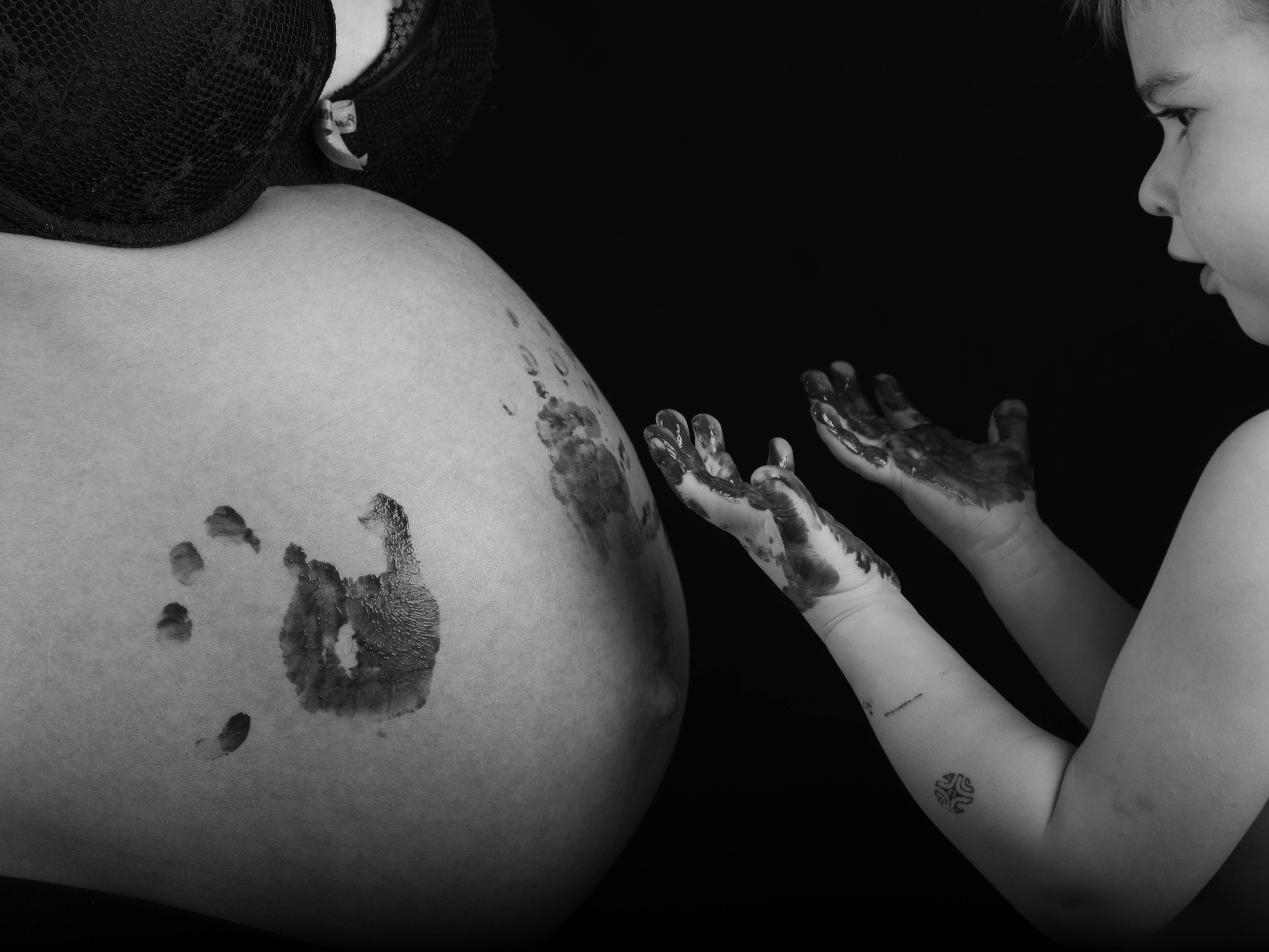 photo noir et blanc d'un ventre de femme enceinte avec les mains de sont enfant en peinture dessus. on voit également la moitié du visage de son enfant qui regarde ses mains barbouillées de peinture