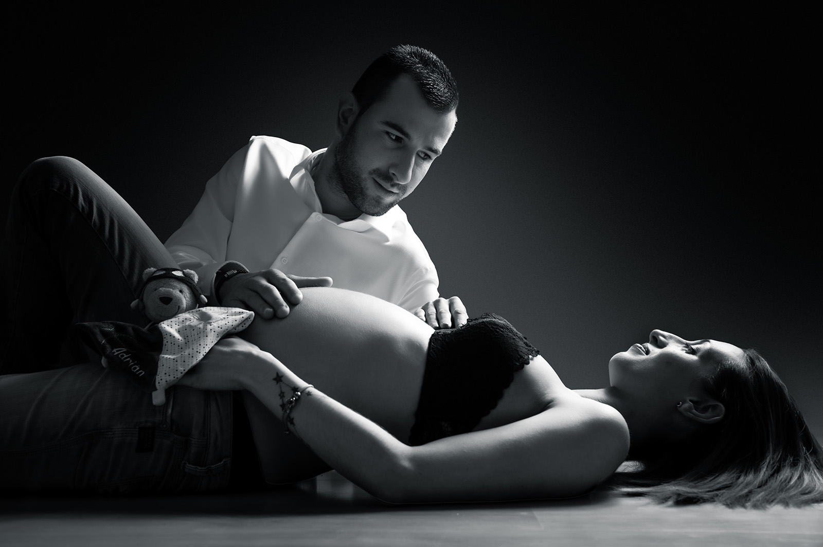 photo noir et blanc d'un couple. ils sont allongés au sol, la femme est enceinte et l'homme a une main posé sur son ventre et la regarde