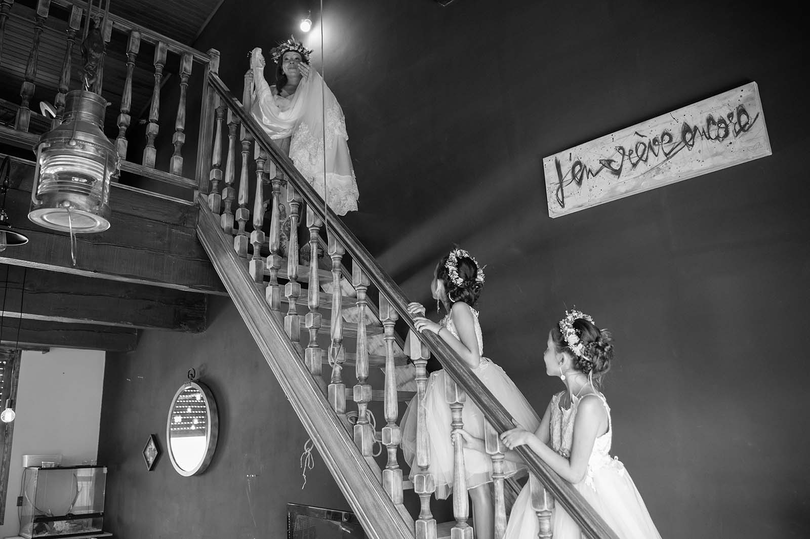 photo noir et blanc d'une dame descendant l'escalier une robe de mariée à la main , deux petites filles sont en bas et la regarde s'avancer