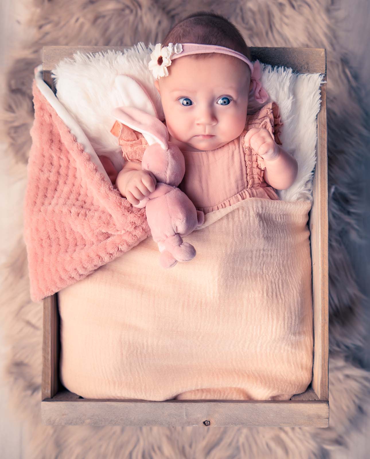 photo d'un bébé positionné sur le dos dans une caisse en bois et tenant son doudou dans la main
