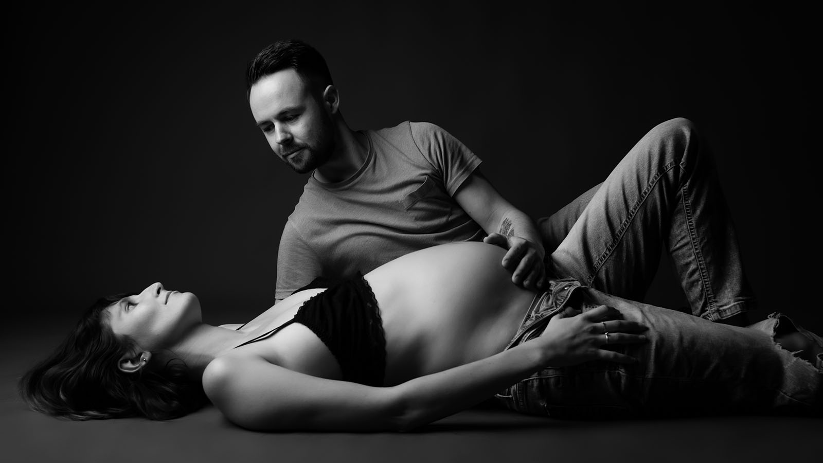 photo noir et blanc d'un couple. ils sont allongés au sol, la femme est enceinte et l'homme a une main posé sur son ventre et la regarde