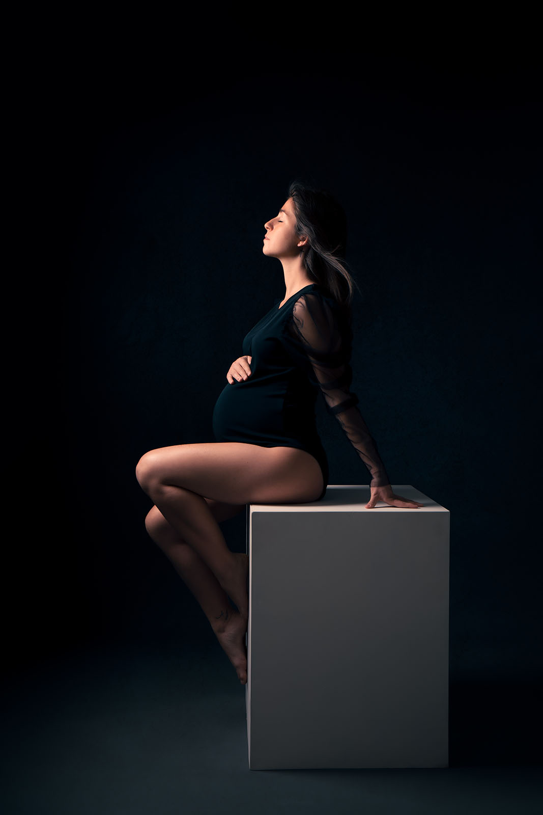 femme enceinte en body noir , elle est assise sur un cube blanc et ses cheveux volent