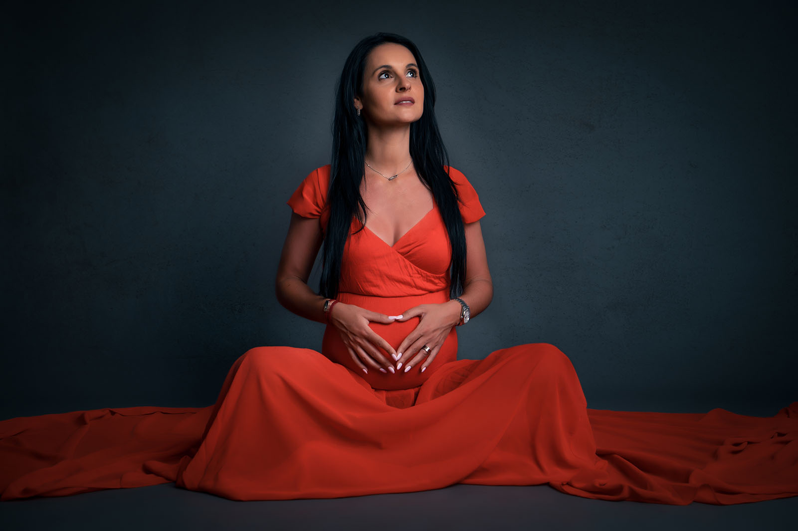 femme enceinte assise au sol vêtue d'une robe rouge, elle est brune a des cheveux longs, son visage est orienté vers le haut et ses mains forment un cœur sur son ventre