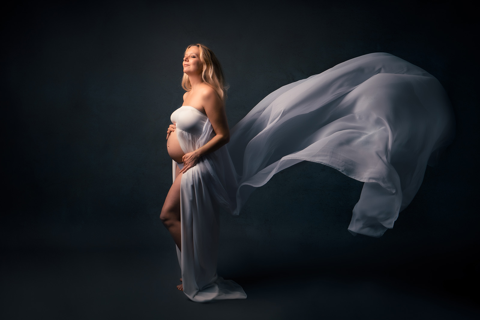 femme enceinte blonde habillée d'une robe en voile blanc dont la partir arrière vole. Elle a les yeux fermés , la tête inclinée vers le haut et elle ferme les yeux