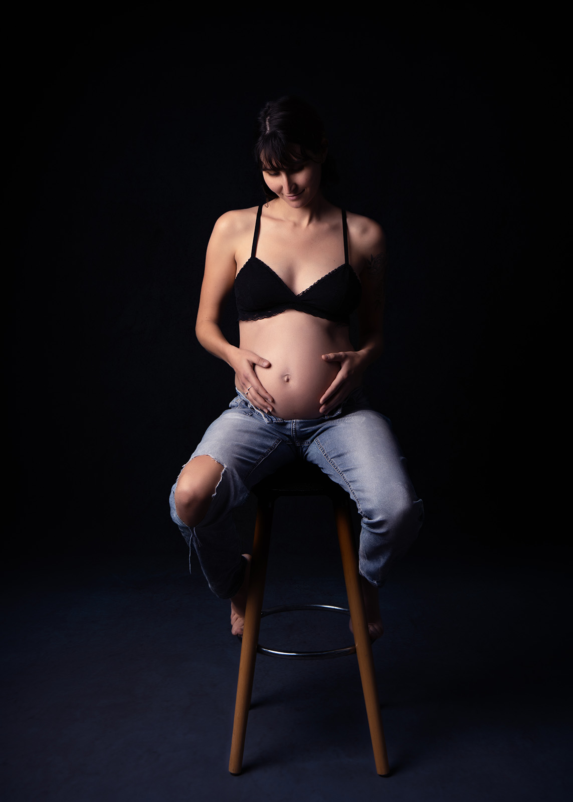 femme enceinte assise sur un tabouret et vêtue d'un soutien gorge noir et d'un jean troué