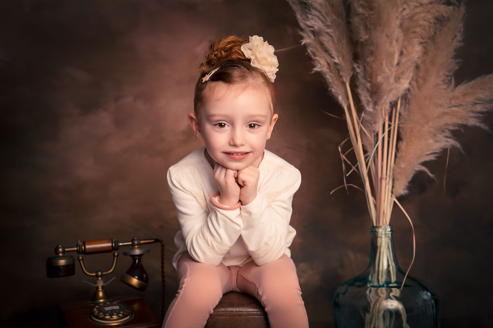 photo d'une petite fille assise se tenant la tête avec les mains dans un décor contenant un téléphone ancien et des fleurs de pampa