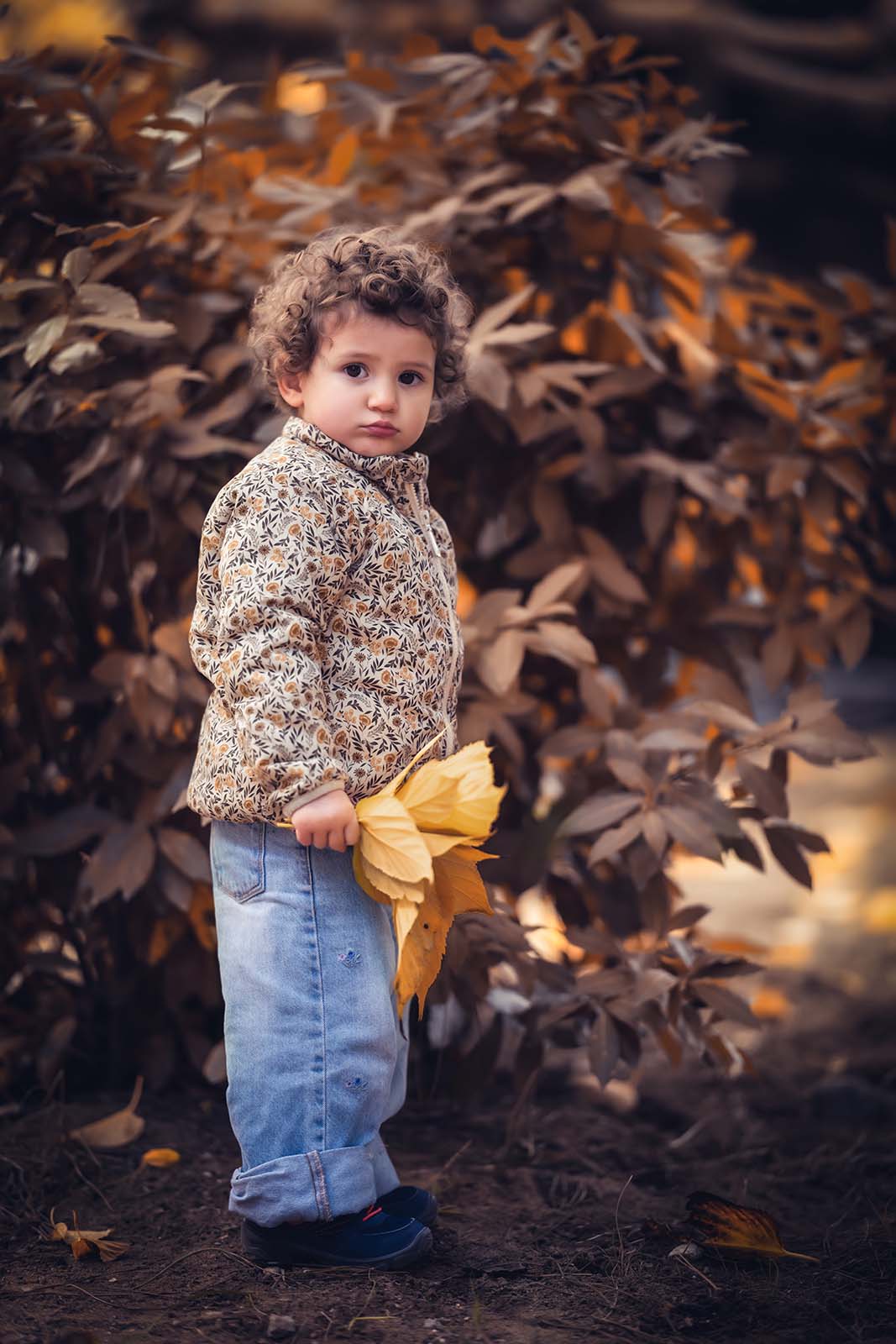 portrait d'une petite fille en extérieur devant des arbres marron et qui tient une grosse feuille jaune à la main