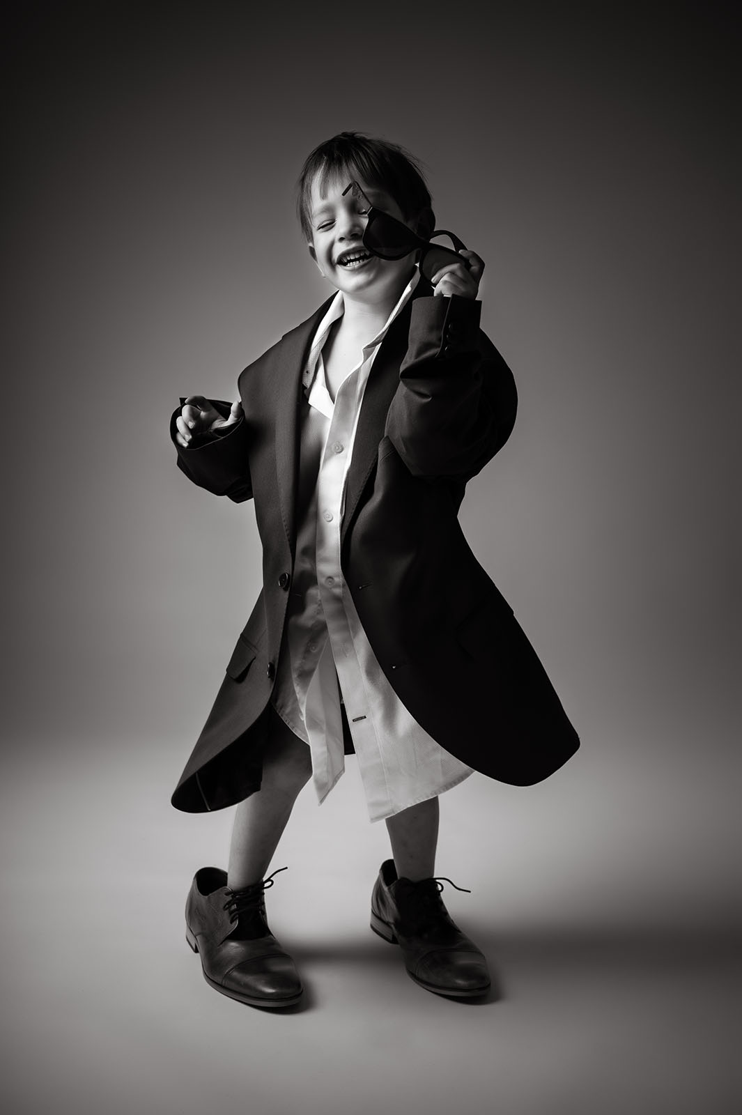 portrait d'un petit garçon en noir et blanc portant une chemise, une veste de costume et des chaussures trop grandes pour lui