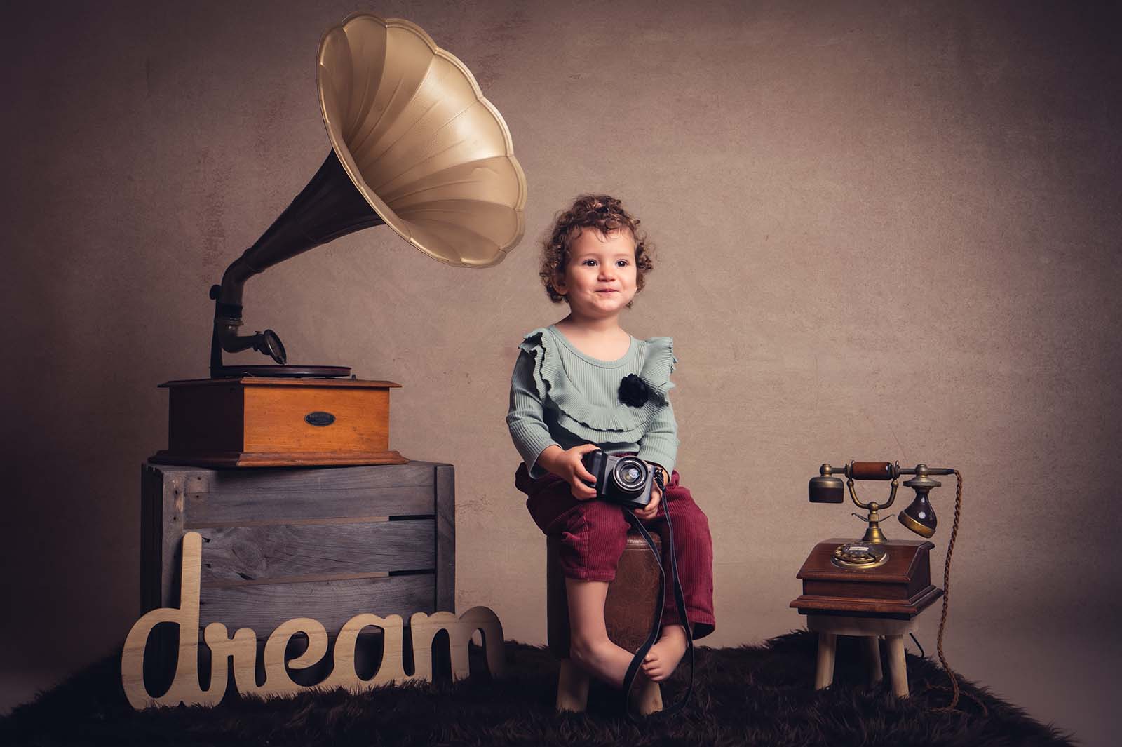 portrait d'une petite fille assise sur un petit tabouret et tenant un appareil photo dans les mains. Ils y a un vieux téléphone et un gramophone sur une caisse en bois comme décor.
