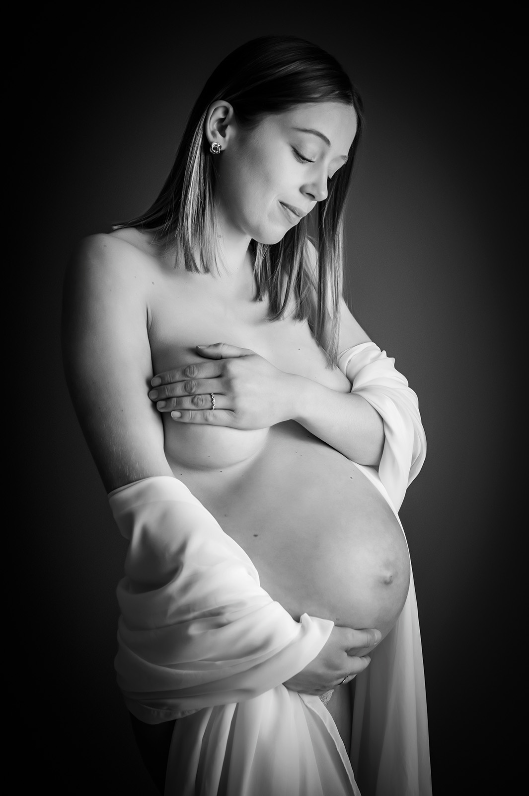 photo noir et blanc d'une femme enceinte habillée d'un voile blanc sur les coudes et se cachant la poitrine avec les mains