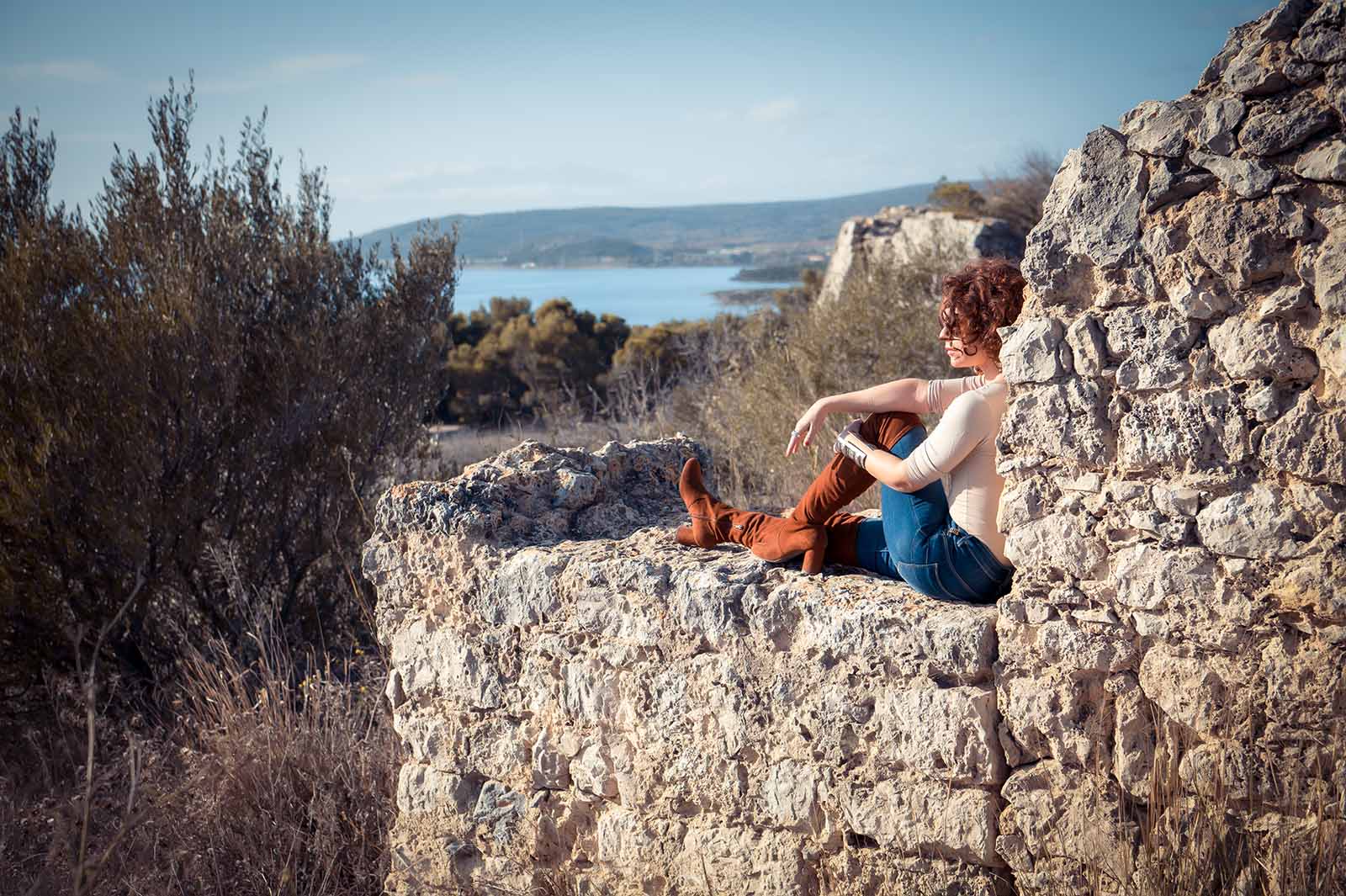jeune femme assise sur un mur de pierres avec une vue sur un étang et des montagnes