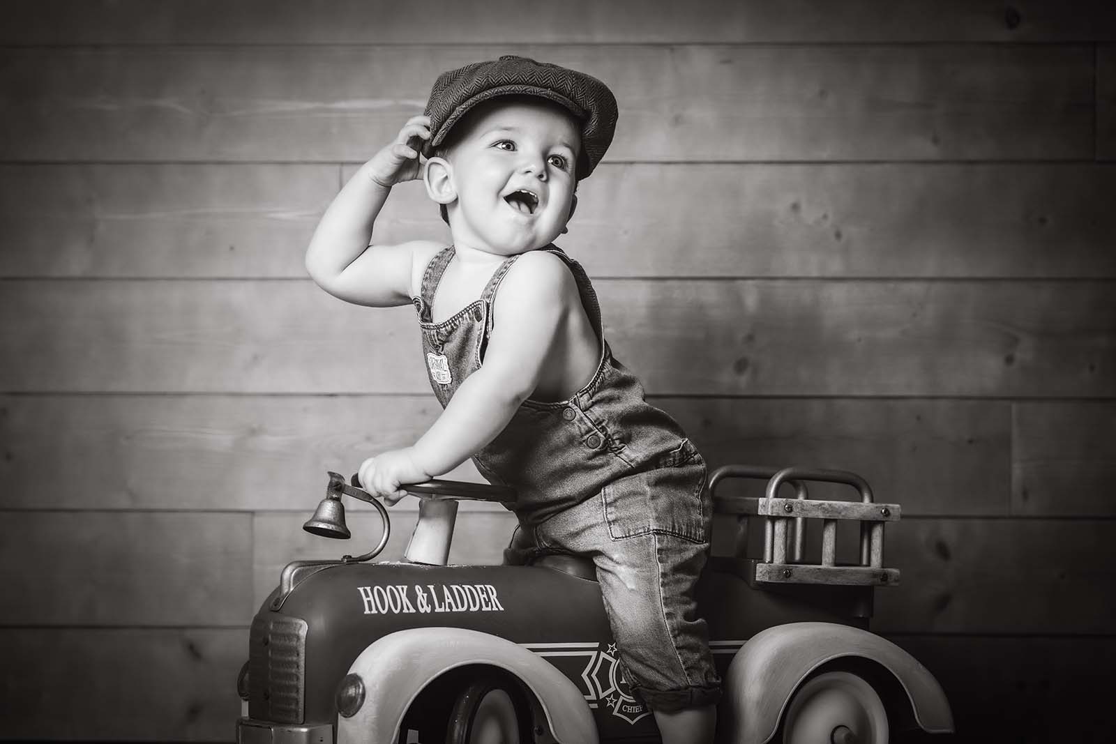 portrait noir et blanc d'un petit garçon sur un petit camion de pompier. Il est habillé en salopette bleue et porte un béret