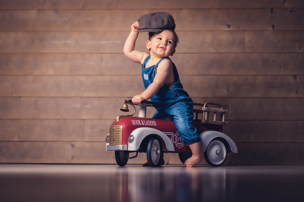 portrait d'un petit garçon sur un petit camion de pompier. Il est habillé en salopette bleue et porte un béret
