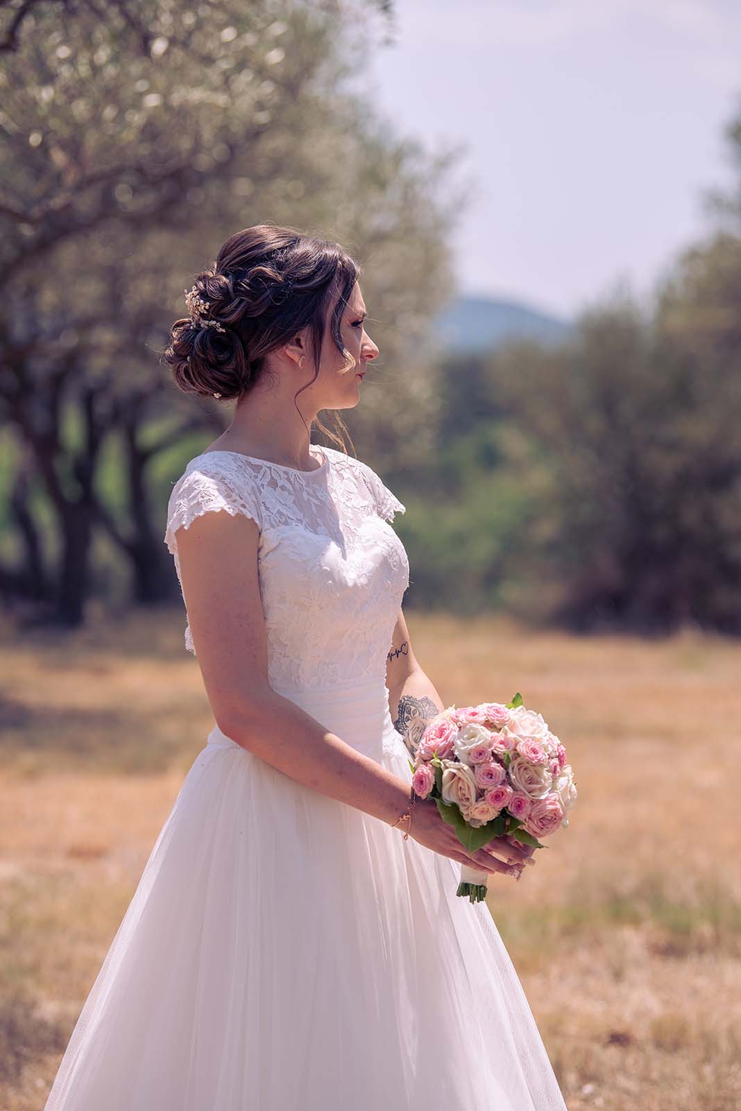 mariée dans un champ d'olivier se tenant de profil bouquet de fleur à la main