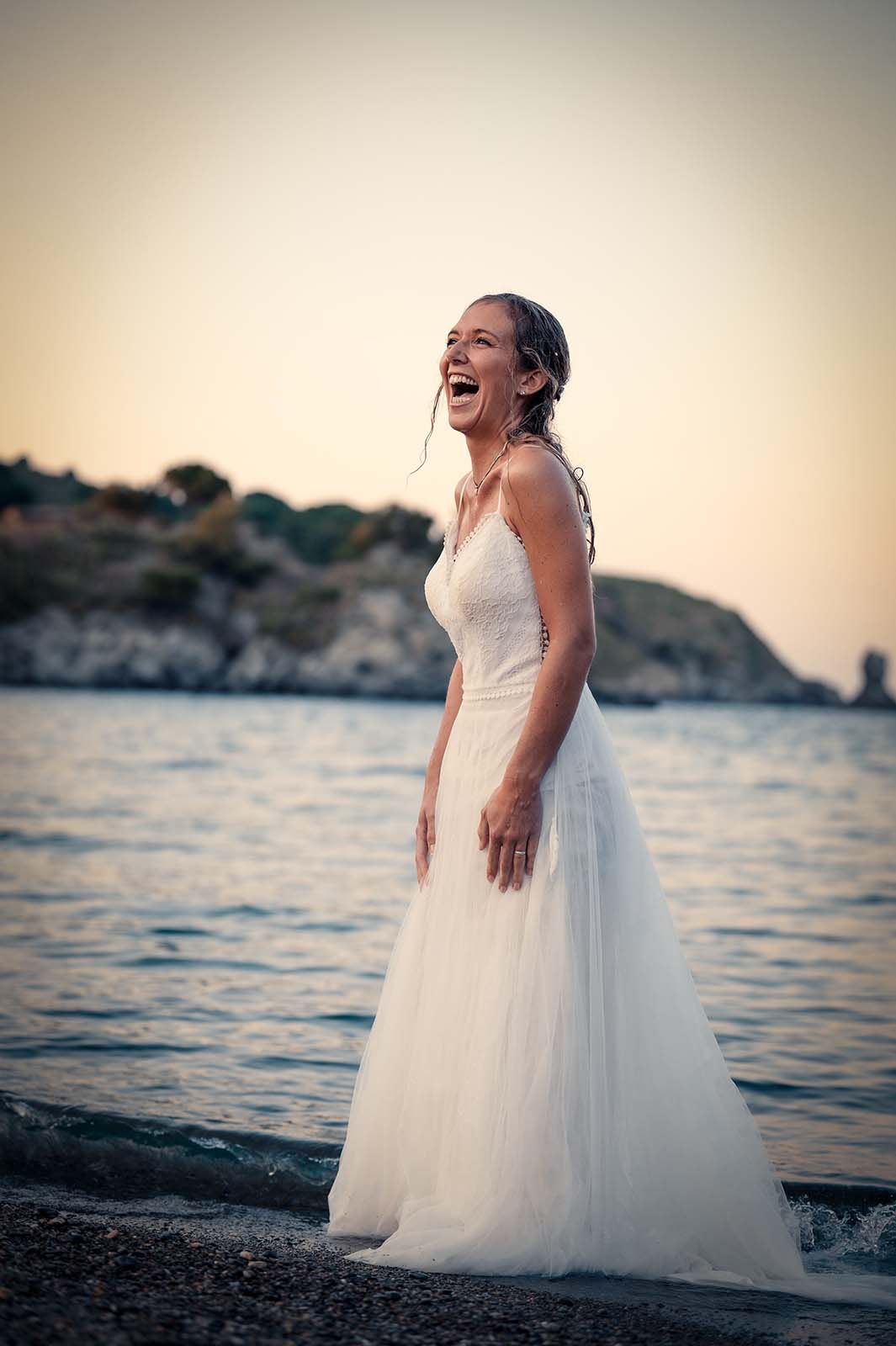 femme blonde en robe de mariée mouillée au bord de l'eau en train d'éclater de rire