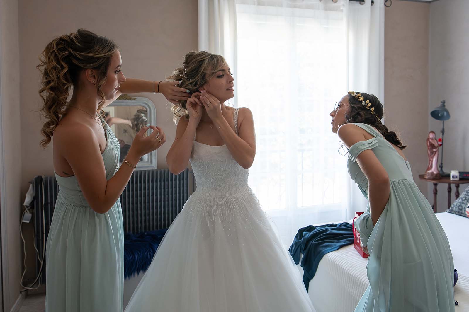 deux jeunes filles et une mariée dans une chambre. La mariée enfile une boucle d'oreille
