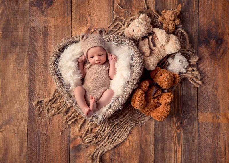 bébé dans un contenant en forme de cœur avec des ours en peluche sur le coté du contenant. Le bébé est habillé en salopette en laine de couleur beige avec un petit bonnet en laine avec des oreilles d'ours