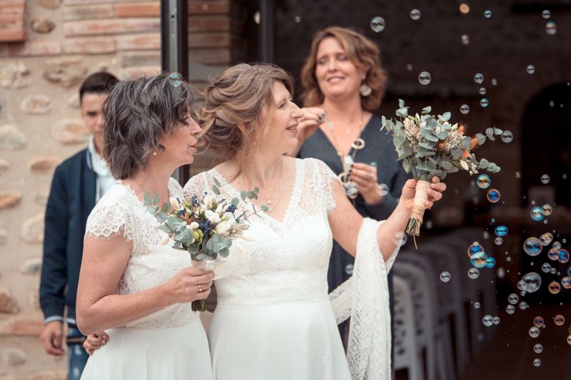 deux femmes habillées en robe de mariées tenant toutes les deux un petit bouquet de fleurs et semblant regarder des bulles qui s'envolent