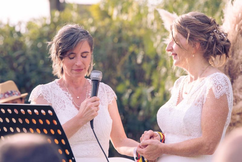 deux femmes habillées en robe de mariée se tenant la main. L'une d'elles tient un micro dans la main