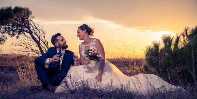 portrait de mariés au coucher de soleil assis au sol en train de se regarder avec un verre et une bouteille de vin à la main