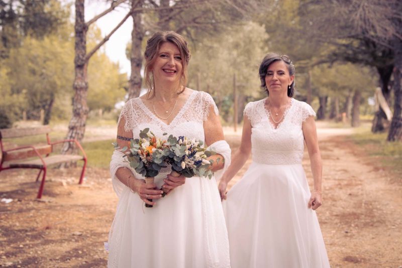 photo de deux femmes en robe de mariées. L'une tient deux bouquets de fleurs et l'autre se situe derrière elle
