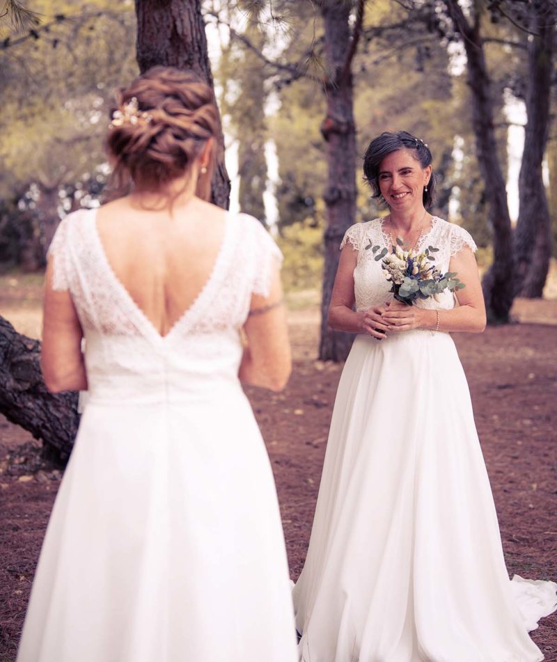 des femmes habillées en robe de mariée. L'un d'elle est de dos et l'autre est face à elle et tient un bouquet de fleurs dans ses mains