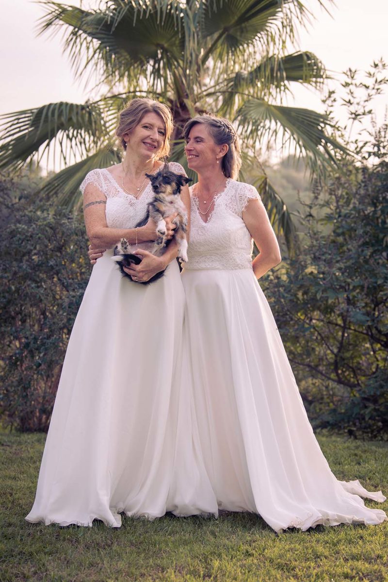 deux femmes habillées en robe de mariée, l'une d'elle tient un petit chien dans les bras