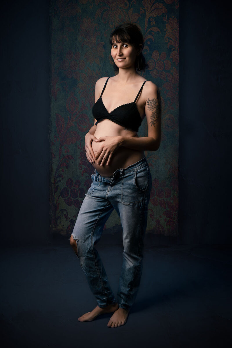 femme enceinte aux cheveux courts brun habillée en soutien gorge noir et jean troué. Ses mains sont positionnées sur son ventre et forme un cœur