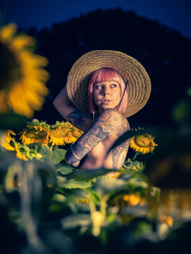 femme tatouée avec des cheveux roses, elle porte un chapeau de paille et se cache la poitrine dans un champ de tournesol