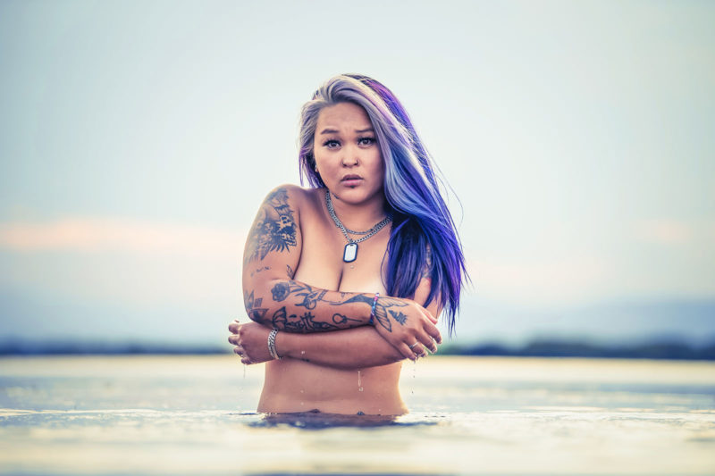 femme d'origine asiatique avec des cheveux longs violet et des tatouages sur le bras et des piercings. Elle est dans l'eau et se cache la poitrine avec les bras