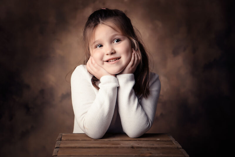 petit fille qui sourit et qui se tient la tête appuyée sur une caisse en bois sur un fond studio marron