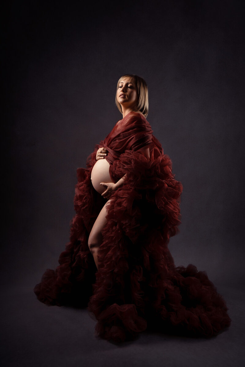 portrait studio d'une femme enceinte de profil portant une longue robe en tulle de couleur bordeaux ouverte sur le ventre