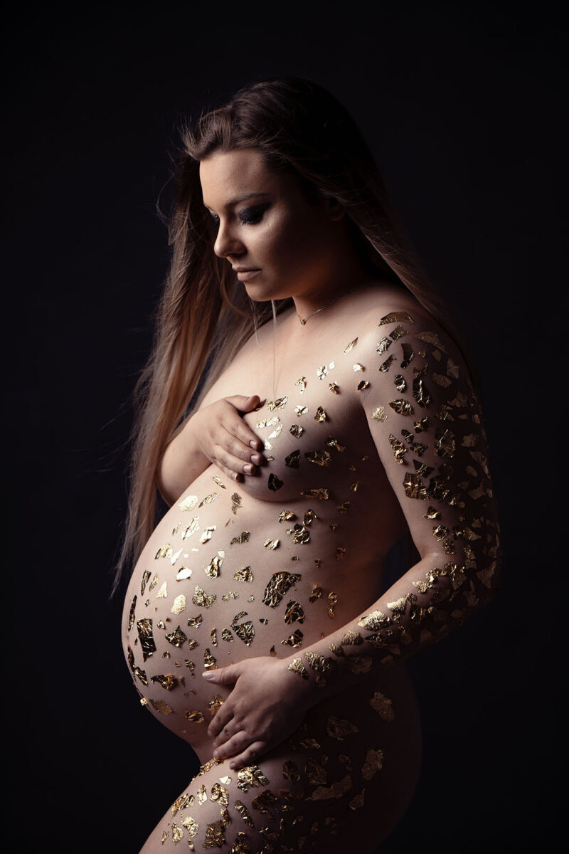 photo d'une femme enceinte blonde nue avec des bouts de feuilles d'or collées sur son corps