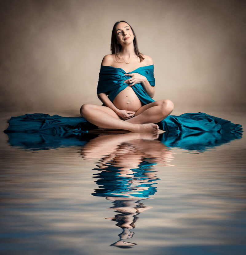 portrait d'une femme enceinte assise en tailleur habillée d'un tissu bleu où l'on voit son reflet dans l'eau