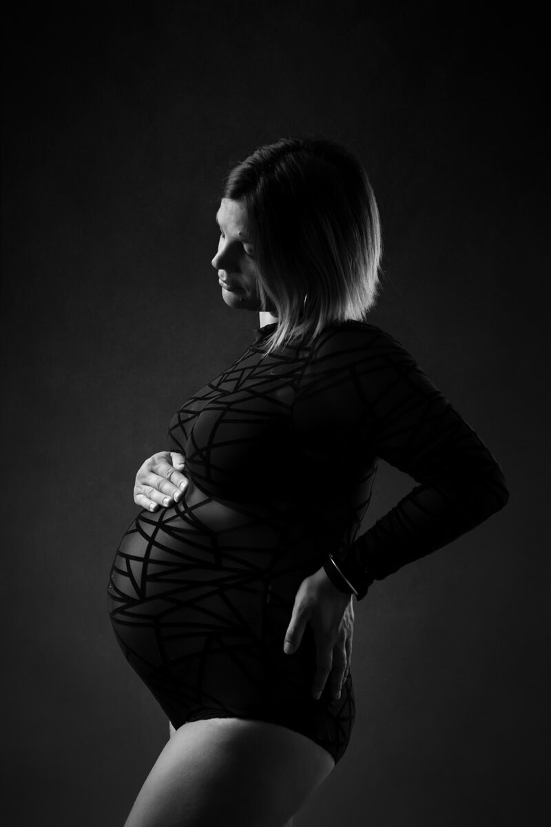 photo noir et blanc d'une femme enceinte en body noir se tenant de profil avec une main sur le hanche et une main sur le ventre
