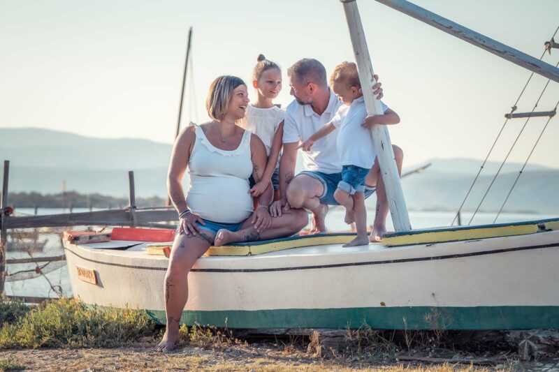 photo d'une famille dans une barque en bas. Ils sont tous habillés en jean et haut blanc, la femme est enceinte et le petit garçon montre le vent de sa maman