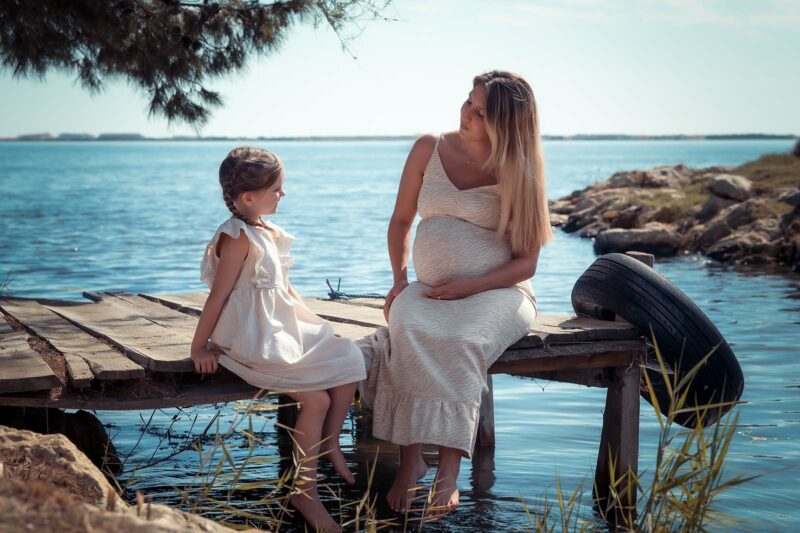 Une femme enceinte et assise sur un ponton au bord de l'eau et elle regarde sa fille qui est assise en face d'elle
