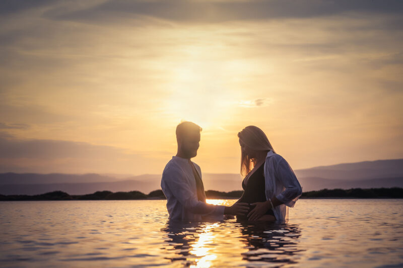 photo d'un couple dans l'eau dont la femme est enceinte au coucher de soleil. Ils sont face à face, l'homme a les mains posées sur le ventre de la femme qui est habillée en maillot de bain noir