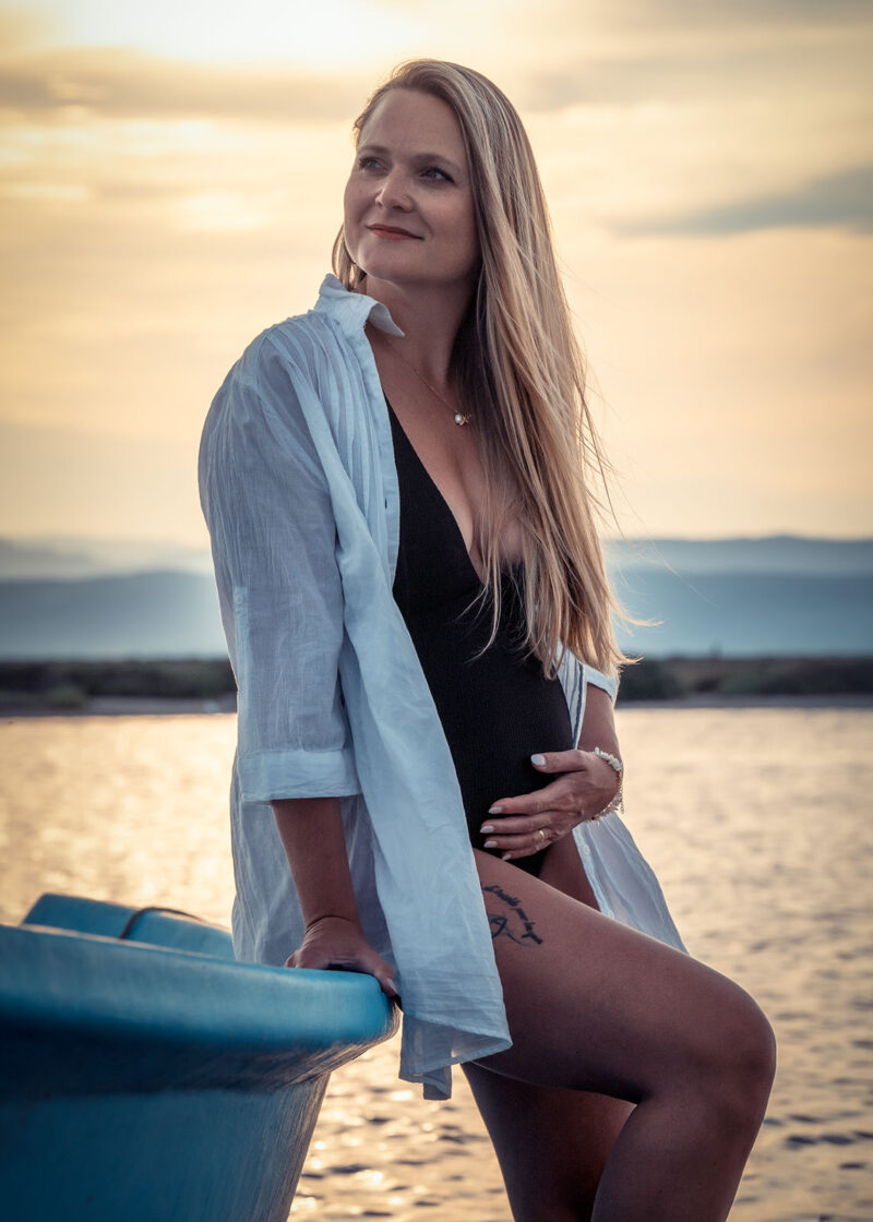 portrait d'une femme enceinte au coucher de soleil habillée en maillot de bain noir une pièce et avec une chemise blanche, elle est assise sur une barque bleu au bord de l'eau