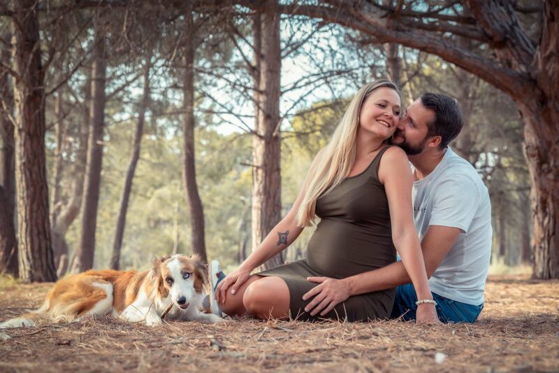 Photo d'un couple assis au sol dans une pinède avec leur chien, la femme est enceinte