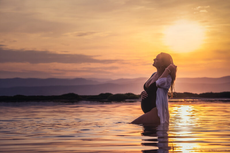 photo d'une femme enceinte habillée en maillot de bain noir une pièce et d'une chemise blanche au coucher de soleil