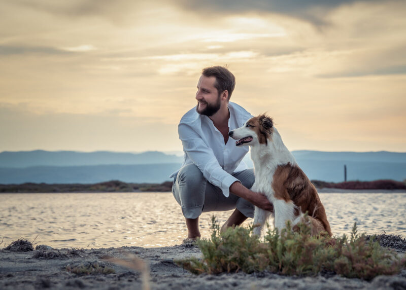 photo d'un homme assis sur le sable accompagné de son chien au coucher de soleil au bord de l'eau