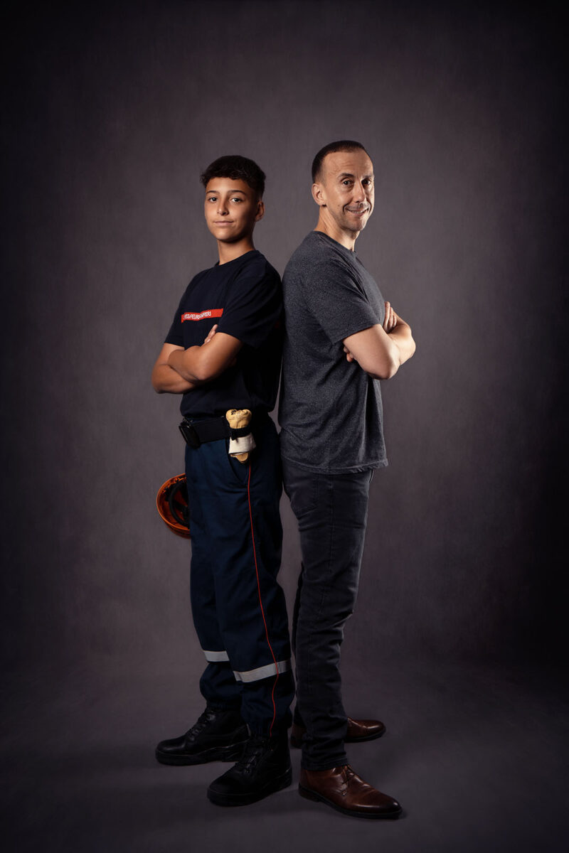 photo studio de deux hommes debout se tenant dos à dos. Le jeune est habillé en tenue de sapeur pompier