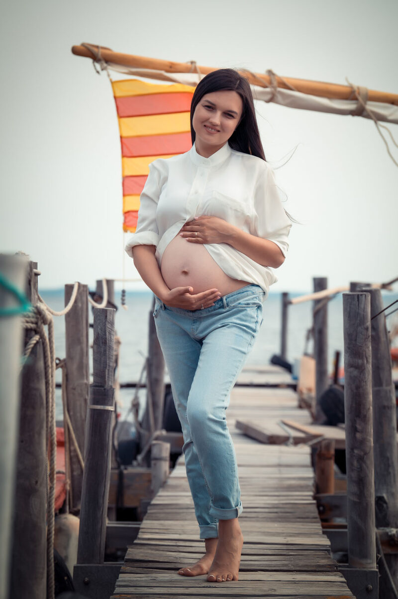 portrait d'une femme enceinte en extérieur. Elle est debout, les mains posées sur le ventre sur un petit ponton en bois