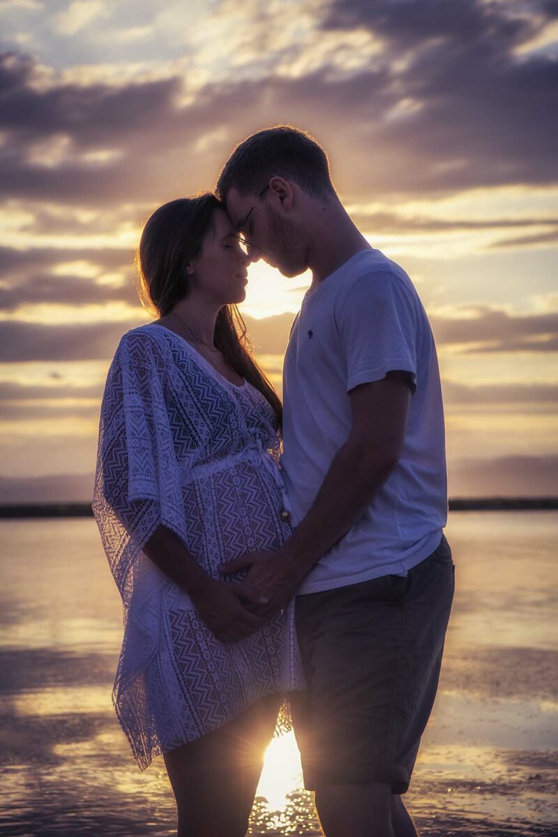 Photo d'un couple où la femme est enceinte, ils ont le front posé l'un contre l'autre au coucher de soleil