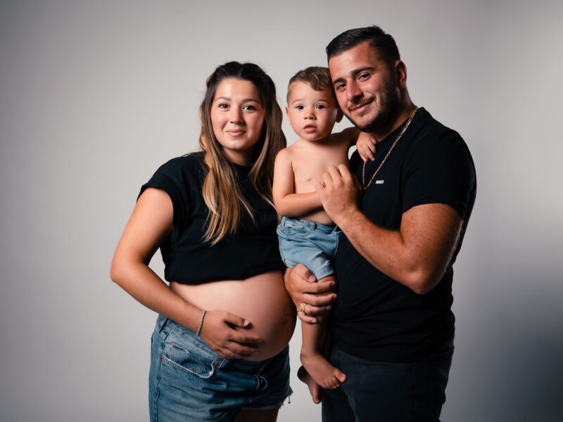 photo studio d'une famille sur fond blanc. La femme est enceinte et le papa tient un petit enfant dans ses bras