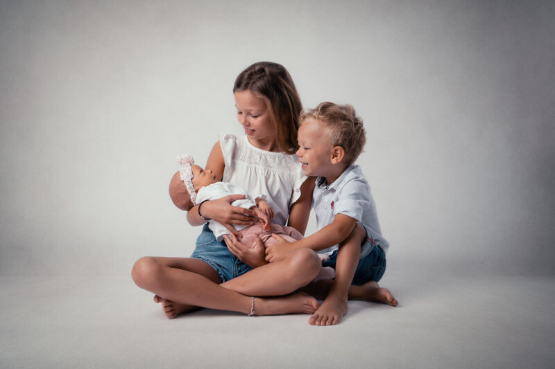 photo studio de deux jeunes enfants assis au sol, la plus grande tient un bébé dans ses bras