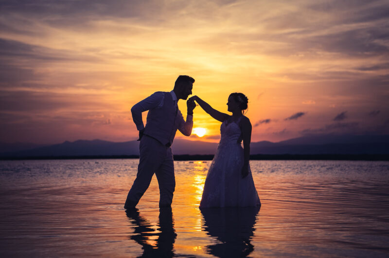 couple de mariés le pieds dans l'eau au coucher de soleil, le marié embrassant la main de la mariée